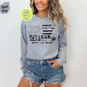 Peut inclure: Un sweat-shirt gris avec un motif de drapeau am&eacute;ricain us&eacute; et le mot "Veteran" imprim&eacute; dessus. Le drapeau est noir et blanc et a une plaque d'identit&eacute; qui pend.
