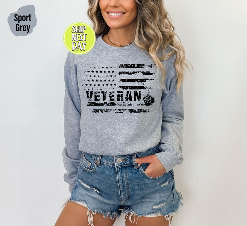 Peut inclure: Un sweat-shirt gris avec un motif de drapeau am&eacute;ricain us&eacute; et le mot "Veteran" imprim&eacute; dessus. Le drapeau est noir et blanc et a une plaque d'identit&eacute; qui pend.