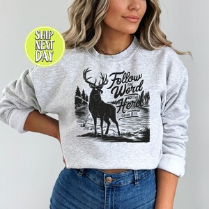 May include: A light gray sweatshirt featuring a black graphic of a deer standing near a lake with trees. The text "Follow the Word Not the Herd" and "Isaiah 8:11" are above the deer. The sweatshirt is a crew neck style.