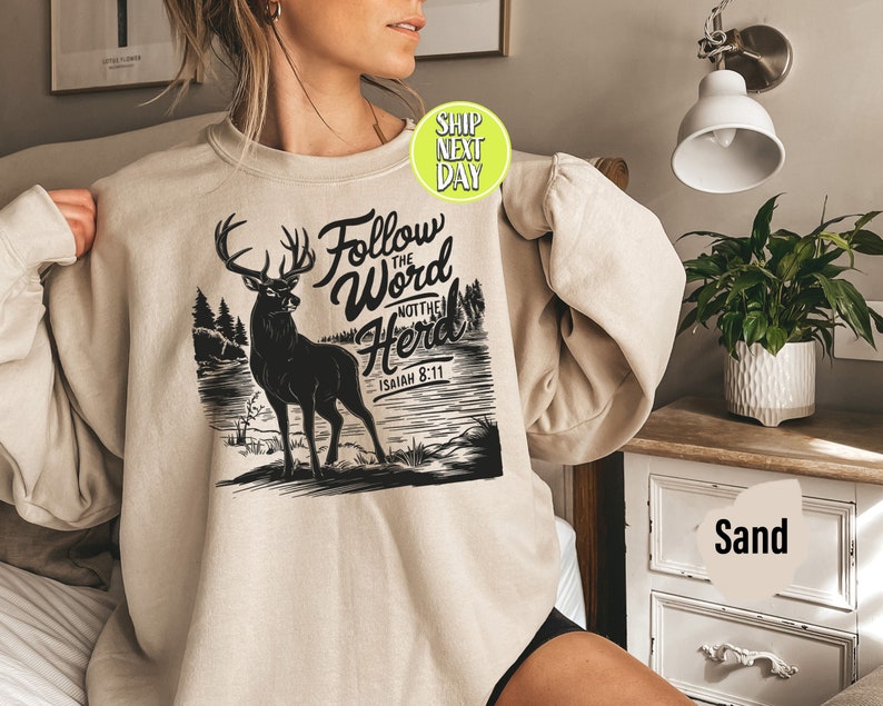 May include: Sand-colored sweatshirt with a black graphic of a deer standing near a lake, with the text "Follow the Word Not the Herd" and "Isaiah 8:11". The sweatshirt has long sleeves and a crew neck.