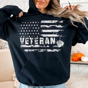 Peut inclure: Sweat-shirt &agrave; capuche noir avec un motif de drapeau am&eacute;ricain d&eacute;lav&eacute; et le mot "Veteran" en blanc. Deux m&eacute;dailles militaires blanches sont sur le c&ocirc;t&eacute; droit du motif.