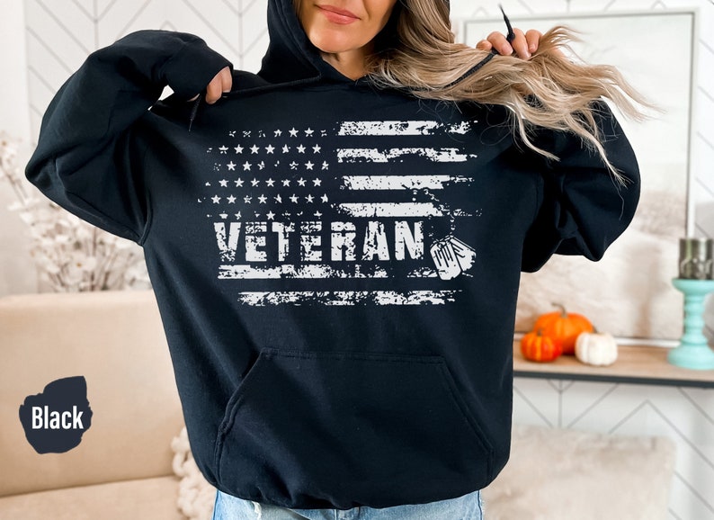 Peut inclure: Sweat-shirt &agrave; capuche noir avec un motif de drapeau am&eacute;ricain d&eacute;lav&eacute; et le mot "Veteran" en blanc. Deux m&eacute;dailles militaires blanches sont sur le c&ocirc;t&eacute; droit du motif.