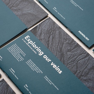 Op de afbeelding: Meerdere rechthoekige donkergroenblauwe brochures met de witte tekst Exploring our veins, A new adventure awaits en de merknaam MONOLISH. Het ontwerp heeft een donkergrijs getextureerd achtergrondpatroon en bevat fotocredits.