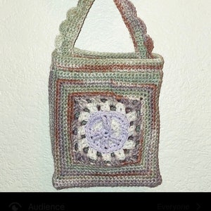 Może przedstawiać: Worek wykonany na szydełku z wzorem znaku pokoju w odcieniach zieleni, brązu, bieli i fioletu. Worek jest zawieszony na drewnianym pierścieniu.