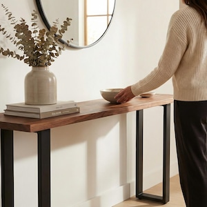 May include: A modern entryway table with a dark wood top and black metal legs. A beige vase with dried foliage sits on the table, along with a stack of books and a decorative bowl. A round mirror hangs on the wall.