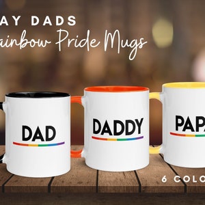 May include: Three white ceramic mugs with black, orange, and yellow rims. Each mug has a different word printed on it: "DAD", "DADDY", and "PAPA". Each word has a rainbow stripe underneath it. The text "6 COLORS" is printed below the mugs.