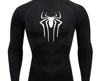 Puede incluir: Camiseta de compresión negra de manga larga con un emblema de araña blanco en el pecho. La camiseta tiene cuello redondo y está diseñada para un ajuste ceñido. El logotipo de la araña es un diseño estilizado y simétrico.