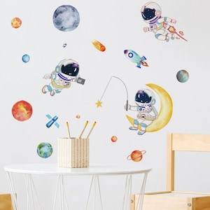 May include: A set of watercolour wall decals depicting astronauts, planets, a rocket, and a crescent moon. The space-themed stickers include colourful planets, a satellite, and a shooting star, ideal for a child's bedroom or playroom.