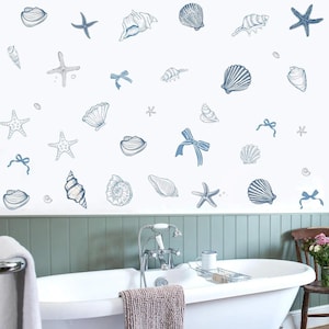 Könnte beinhalten: Eine weiße Wand mit blauen Illustrationen von Muscheln und Seesternen. Das Design umfasst verschiedene Muscheltypen, Seesterne und Schleifen, die ein Küsten- oder nautisches Thema schaffen. Eine weiße Badewanne und Badezimmerarmaturen sind ebenfalls zu sehen.