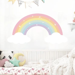 Peut inclure: Un autocollant mural en forme d'arc-en-ciel pastel avec des nuages blancs et des étoiles scintillantes. L'arc-en-ciel présente des bandes de rose, de jaune, de bleu et de violet. Il est placé au-dessus d'un lit bébé blanc.