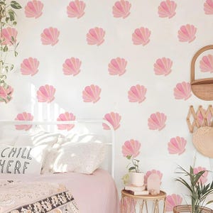 May include: A bedroom featuring a white bed frame, pink bedding, and a wall adorned with pink seashell decals. A pillow on the bed displays the text "CHILL HERE". A small table holds books and a plant. A mirror and hat rack are on the wall.