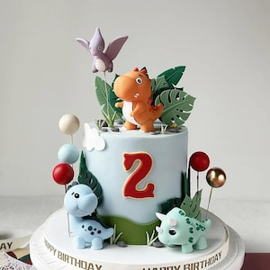 May include: A light blue birthday cake decorated with dinosaur figurines, green leaves, and red number 2. The cake has a white base with the words "Happy Birthday" around the edge. Colorful cake pops and a purple pterodactyl complete the festive design.