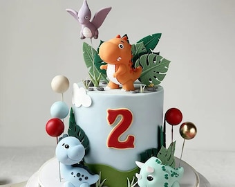 May include: A light blue birthday cake decorated with dinosaur figurines, green leaves, and red number 2. The cake has a white base with the words "Happy Birthday" around the edge. Colorful cake pops and a purple pterodactyl complete the festive design.