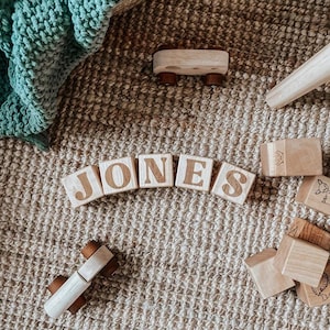 May include: Wooden blocks spelling "JONES" are arranged on a textured surface, alongside other wooden toys. The blocks are light-colored with brown lettering. A small wooden car and other blocks with animal illustrations are also present.