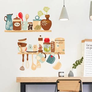 Op de afbeelding: Een muursticker met twee planken met illustraties met keukenthema. De bovenste plank toont een theepot, potten met het opschrift "TEA" en lepels. De onderste plank toont mokken, potten en keukengerei. Het kleurenpalet omvat blauw, bruin en beige.