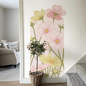 May include: A wall decal featuring a floral design with large pink and yellow cosmos flowers. The flowers have detailed line art and are set against a light pink background. A small tree in a woven basket is in the foreground.