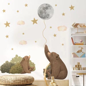 May include: Wall stickers showing two brown bears, one reaching for a grey moon balloon and the other in a green bush. Gold stars and pink clouds decorate the white background. A whimsical nursery theme.