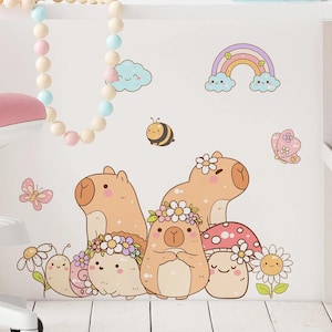 Pode incluir: Um conjunto de decalques de parede caprichosos com capivaras de desenho animado, um cogumelo, uma abelha, uma borboleta, um arco-íris e nuvens. As capivaras são adornadas com coroas de flores. A paleta de cores inclui tons de castanho, rosa, amarelo e azul.