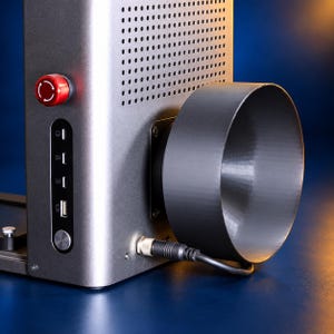 May include: A metallic gray industrial machine featuring a perforated ventilation panel, a prominent red emergency stop button, and a vertical control interface with multiple ports. A large, dark, funnel-shaped attachment is mounted on the side.