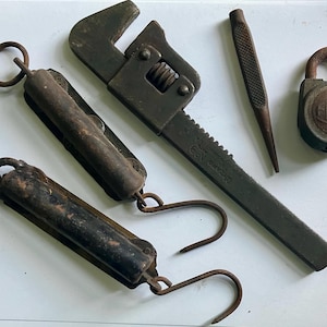 Puede incluir: Un conjunto de herramientas antiguas de metal oxidado, que incluye dos básculas colgantes, una llave inglesa, un punzón y un candado. El candado tiene la palabra "GRAIN". Las herramientas muestran signos de desgaste.