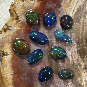May include: A collection of nine polished black opal gemstones with iridescent flashes of green, blue, and red. The stones are oval, pear, and teardrop shaped, displaying a dark base color with vibrant, multi-colored play-of-color throughout.