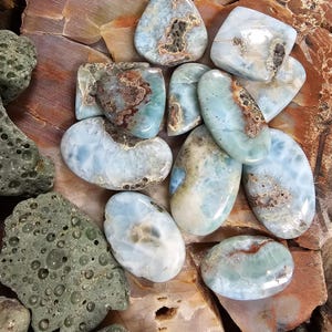 May include: A collection of polished gemstones in various shapes and sizes. The stones feature a mix of blue, white, and brown hues, with unique patterns and textures. Some stones are oval, while others are teardrop or irregular shapes.