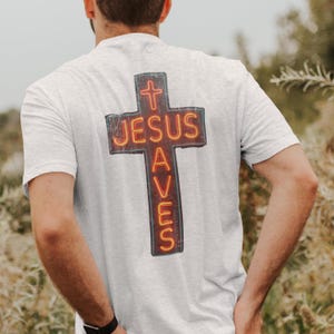 May include: A light gray t-shirt featuring a cross design with the words "JESUS SAVES" in a neon-style font. The cross is outlined in a dark color with a red and orange glow.