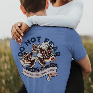 May include: A light blue t-shirt featuring a graphic of two birds with red, brown, and white details, and the words "DO NOT FEAR" above them. Below the birds is a banner that reads "YOU ARE MORE VALUABLE THAN MANY SPARROWS" with a Bible verse reference.