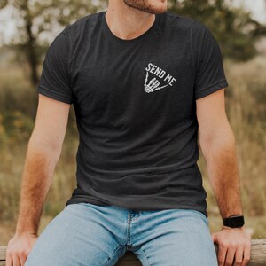 Puede incluir: Camiseta gris oscuro con el texto "SEND ME" sobre un gesto de mano de esqueleto blanco. La camiseta de manga corta se usa con jeans azules. La persona est&aacute; sentada afuera en una valla de madera.