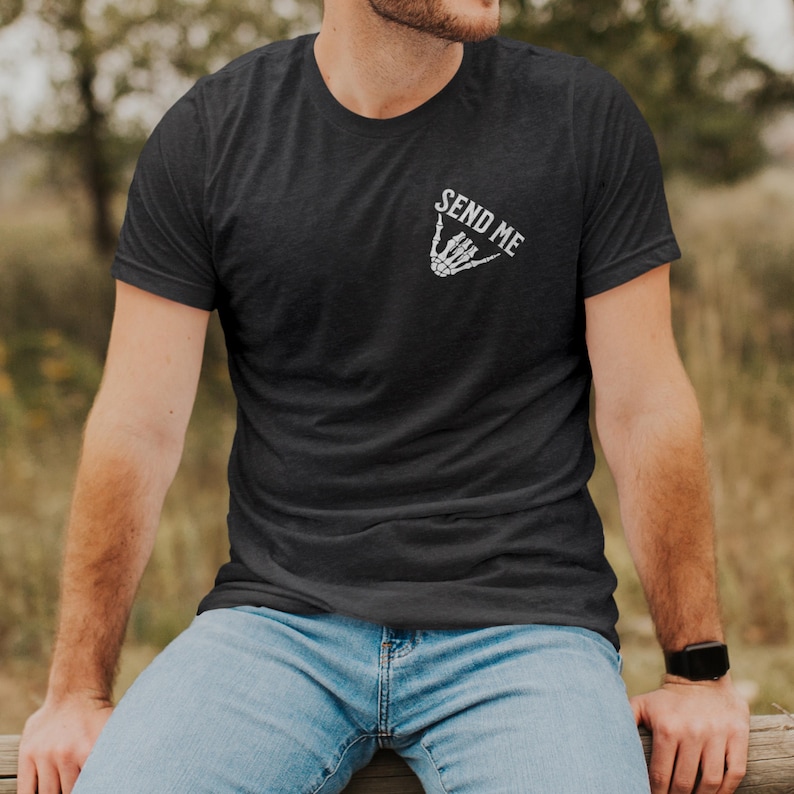 Puede incluir: Camiseta gris oscuro con el texto "SEND ME" sobre un gesto de mano de esqueleto blanco. La camiseta de manga corta se usa con jeans azules. La persona est&aacute; sentada afuera en una valla de madera.
