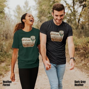 Puede incluir: Una pareja lleva camisetas a juego con un dise&ntilde;o de estilo vintage. Las camisetas presentan un cartel marr&oacute;n con el texto "Bienvenido a nuestra aventura Luke & Sarah 12.12.2025". El hombre lleva una camiseta de color gris oscuro jaspeado y la mujer lleva una camiseta de color verde jaspeado.