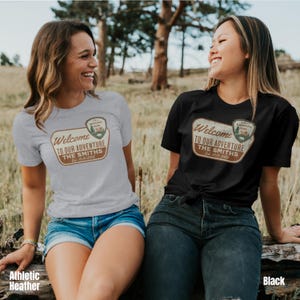 Puede incluir: Dos mujeres vistiendo camisetas con un logotipo de parque nacional de estilo vintage y el texto "Bienvenido a nuestra aventura The Smiths Est. 2025". La mujer de la izquierda lleva una camiseta gris jaspeada y la mujer de la derecha lleva una camiseta negra.