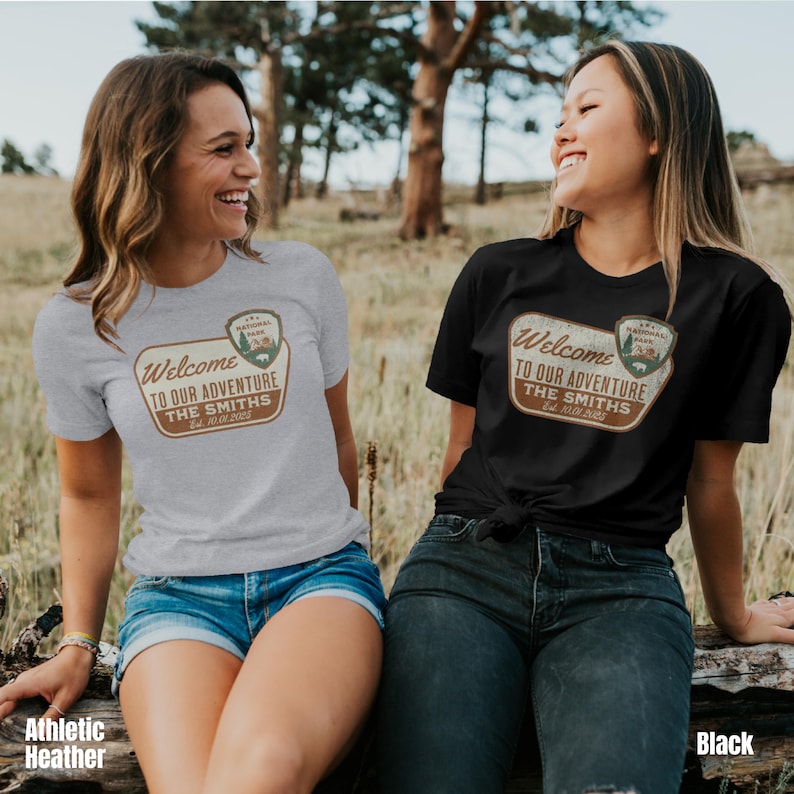 Puede incluir: Dos mujeres vistiendo camisetas con un logotipo de parque nacional de estilo vintage y el texto "Bienvenido a nuestra aventura The Smiths Est. 2025". La mujer de la izquierda lleva una camiseta gris jaspeada y la mujer de la derecha lleva una camiseta negra.