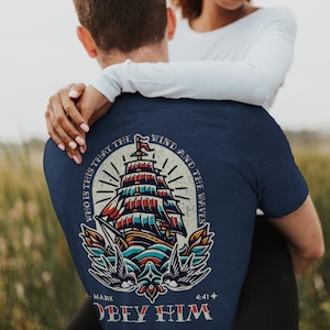 May include: Navy blue t-shirt featuring a detailed illustration of a sailing ship with red, blue, and white accents. The design includes the text "WHO IS THIS THAT THE WIND AND THE WAVES OBEY HIM" and "MARK 4:41".