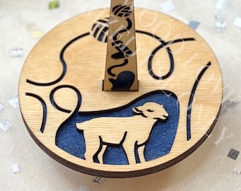 Pode incluir: Um novelo de madeira com uma base circular e um suporte central. A base apresenta um design recortado com uma silhueta de ovelha e um fundo azul. O suporte tem um padrão decorativo. Um anel de metal é fixado no topo.