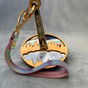 May include: A wooden keychain stand with a gold-coloured clasp and a multi-coloured leather strap. The stand features a laser-cut design of a winter landscape with trees and a lake. The strap has a purple and blue iridescent finish.