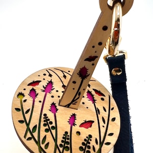 May include: A wooden keychain with a floral design. The keychain features a round base with a floral pattern and a long, thin wooden handle with a gold ring at the top. The handle is attached to a blue leather strap.