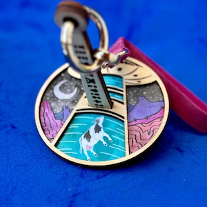 May include: A wooden keychain with a circular pendant featuring a cow leaping over a turquoise body of water. The design includes a night sky with a crescent moon and mountains. A pink ribbon and a metal key ring are attached.