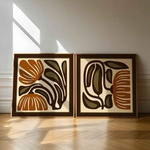May include: Two framed abstract floral art prints. Each print features a brown frame, a cream-colored background, and stylized flowers in shades of brown, orange, and olive green. The artwork is displayed against a white wall.