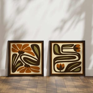 May include: Two framed art prints with abstract floral designs. The artwork features orange, brown, and olive green shapes on a cream background, set within dark brown frames. The prints are displayed on a wooden floor against a white wall.