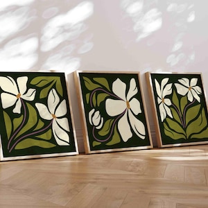 May include: Three framed floral art prints. Each print features a large white flower with a yellow centre and purple stem against a dark green background. The frames are light wood, and the art leans against a white wall.