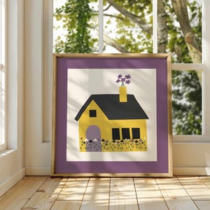 Op de afbeelding: Een ingelijste kunstdruk met een geel huis met een zwart dak en schoorsteen. Het huis heeft een paarse deur en is omgeven door paarse en gele bloemen. Het kunstwerk staat tegen een witte achtergrond met een paarse rand.