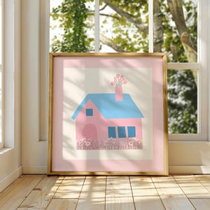 May include: A framed art print depicting a pink house with a blue roof and chimney. The house has three blue windows and a small flower garden at the base. The artwork is set against a cream background and framed in a gold-coloured frame.