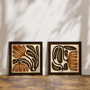 May include: Two framed art prints with abstract floral designs. Each print has a square format with a dark brown frame. The artwork features stylized flowers in shades of brown, olive green, and cream, creating a modern aesthetic.