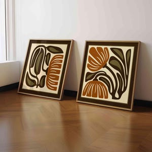 May include: Two framed art prints with abstract floral designs. The artwork features brown, olive green, and cream colors. The frames are a dark brown color. The prints are leaning against a white wall on a wooden floor.