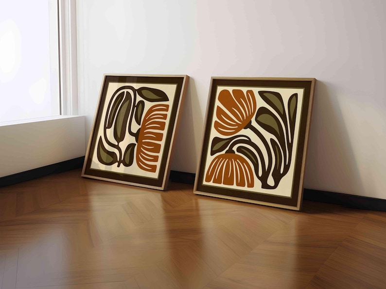 May include: Two framed art prints with abstract floral designs. The artwork features brown, olive green, and cream colors. The frames are a dark brown color. The prints are leaning against a white wall on a wooden floor.