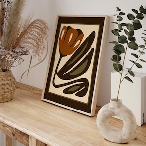 K&ouml;nnte beinhalten: Gerahmter Kunstdruck mit abstraktem Blumendesign in Braun-, Olivgr&uuml;n- und Cremet&ouml;nen. Das Kunstwerk steht auf einem Holztisch. Eine Vase mit kreisf&ouml;rmiger &Ouml;ffnung und getrockneten Pflanzen sind ebenfalls zu sehen.