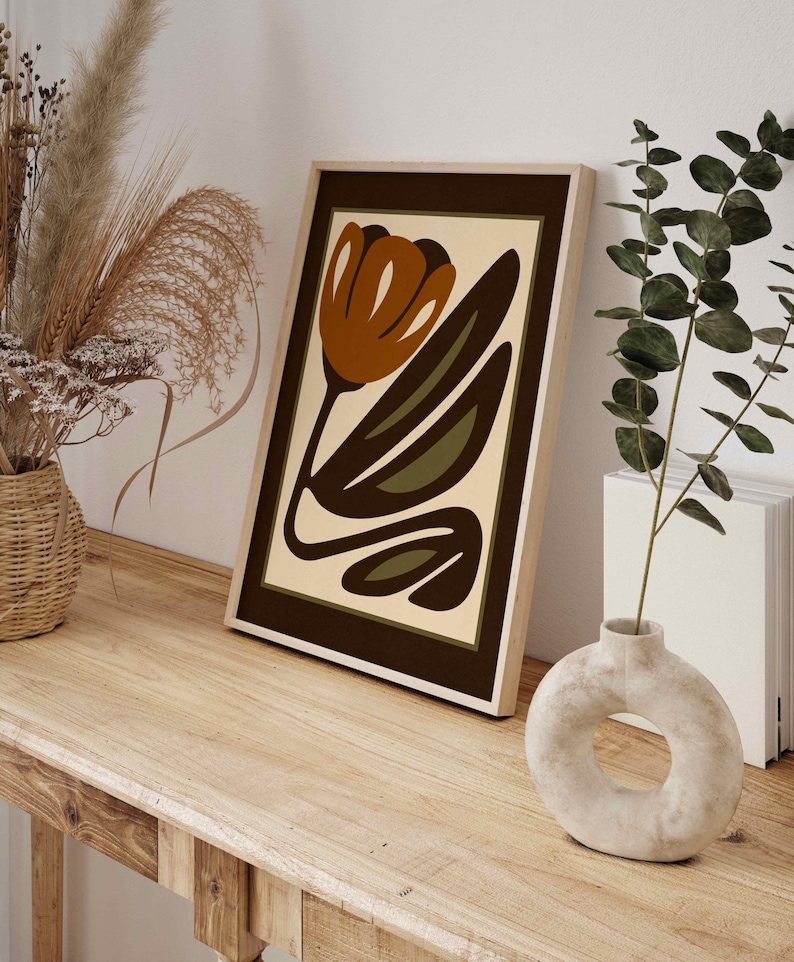 K&ouml;nnte beinhalten: Gerahmter Kunstdruck mit abstraktem Blumendesign in Braun-, Olivgr&uuml;n- und Cremet&ouml;nen. Das Kunstwerk steht auf einem Holztisch. Eine Vase mit kreisf&ouml;rmiger &Ouml;ffnung und getrockneten Pflanzen sind ebenfalls zu sehen.