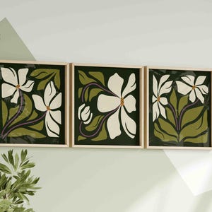 May include: A triptych of framed floral prints. Each print showcases a white flower with a yellow centre, set against a dark green backdrop with olive green leaves and purple stems. The frames are light wood, creating a coordinated wall art display.