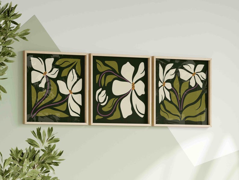 May include: A triptych of framed floral prints. Each print showcases a white flower with a yellow centre, set against a dark green backdrop with olive green leaves and purple stems. The frames are light wood, creating a coordinated wall art display.